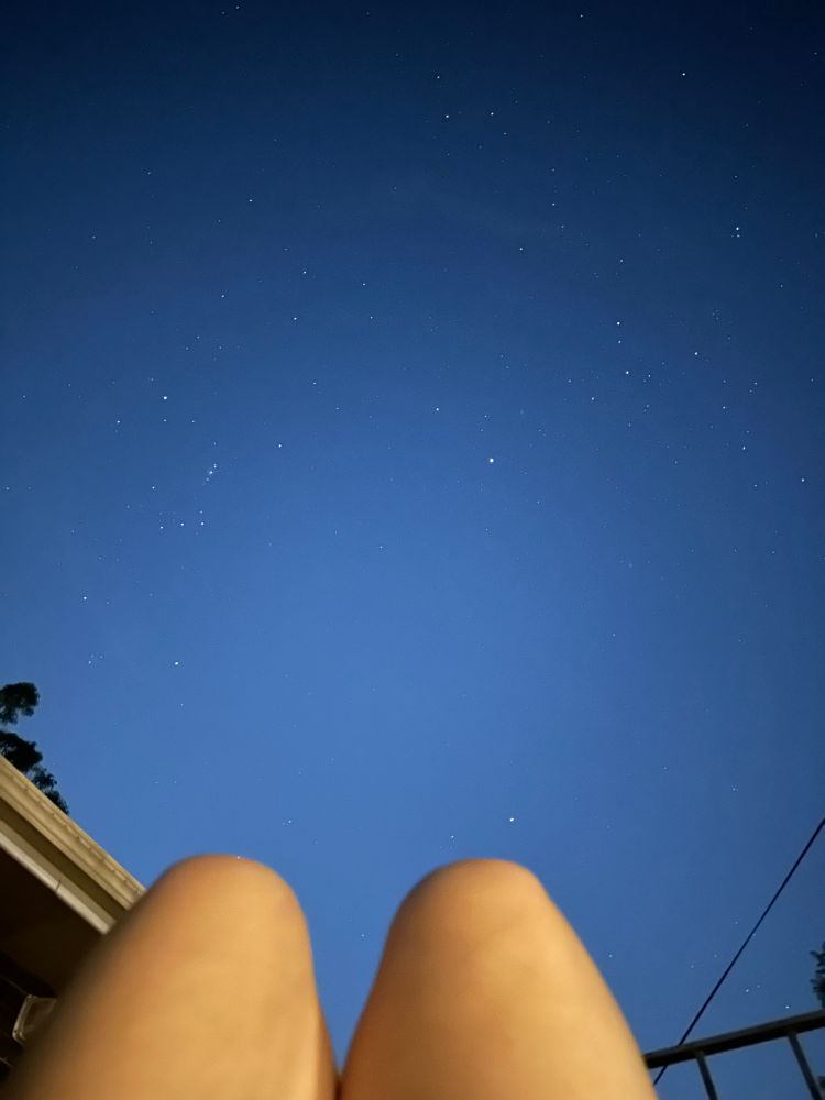 A woman’s knees bent up at the bottom of the image beneath a starry night sky featuring both Orion and Mars