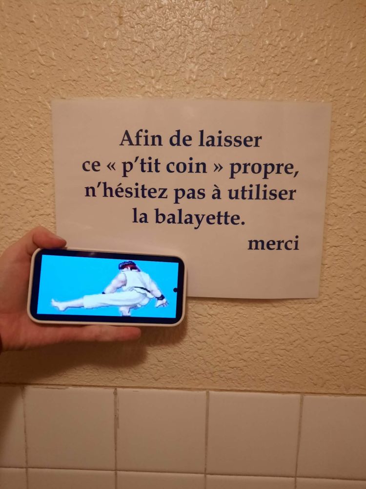 Message dans les toilettes "afin de laisser ce p'tit coin propre, n'hésitez pas à utiliser la balayette. Merci"
À côté une main qui tient un téléphone et dessus une image de ryu qui fait un bas gros pied 