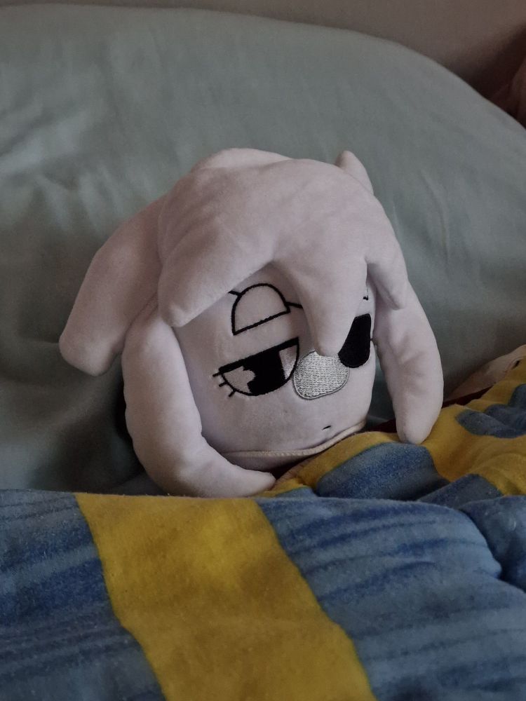 A photo of a Siffrin plushie tucked in a bed, under a blue blanket with yellow stripes and a light blue pillow.