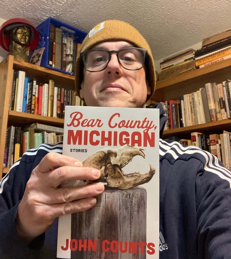 In John’s selfie, he’s holding up a copy of his new book, “Bear County, Michigan.”