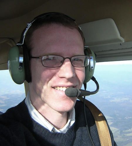 A young man with a light green headset and blue sweater in the left front seat of a small aircraft.