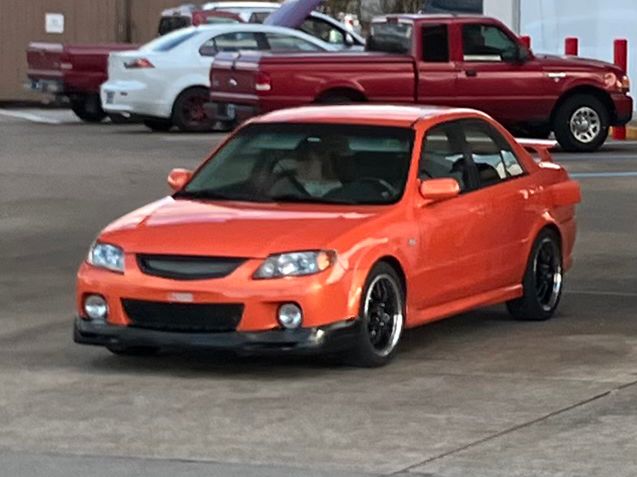 Orange BJ Mazda Protege with some aftermarket modifications including black wheels with polished lip, rally-style foglights and a body kit.