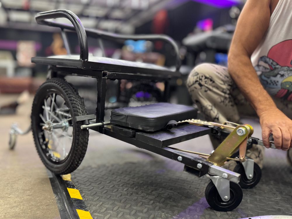 A detailed, low-angle photograph showcases a black metal dog cart, resting on a black, diamond-plate rubber floor. The cart is customized for pony play: built low to the ground, featuring two large, knobby-tired wheels on either side and smaller caster wheels at the front and rear for agility. It has a small, black padded seat and a lower, longer padded platform for kneeling, complete with a strap