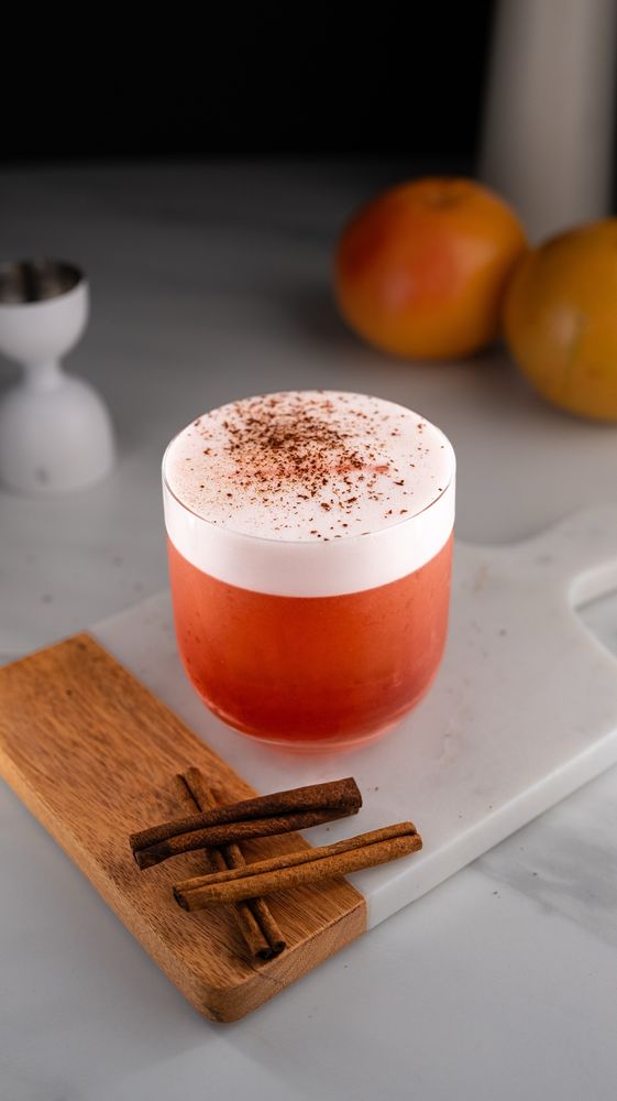 A vibrant cocktail in a rocks glass with a creamy foam layer topped with grated cinnamon. The drink has a reddish-pink hue and sits on a marble surface, accompanied by a wooden board holding a few cinnamon sticks. The background features blurred oranges, creating a warm, inviting scene.