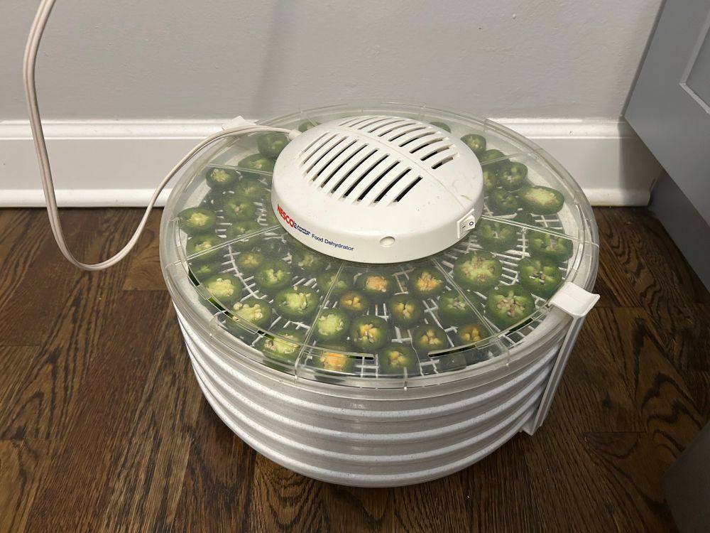 A round food dehydrator with a clear plastic top showing stacked trays of jalapeño slices. 