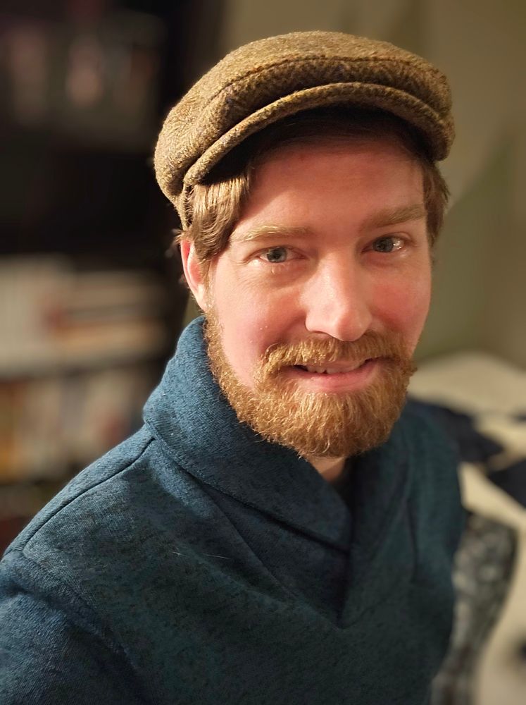 The bearded poster smiling at the camera wearing an emerald sweater and green newsie cap.
