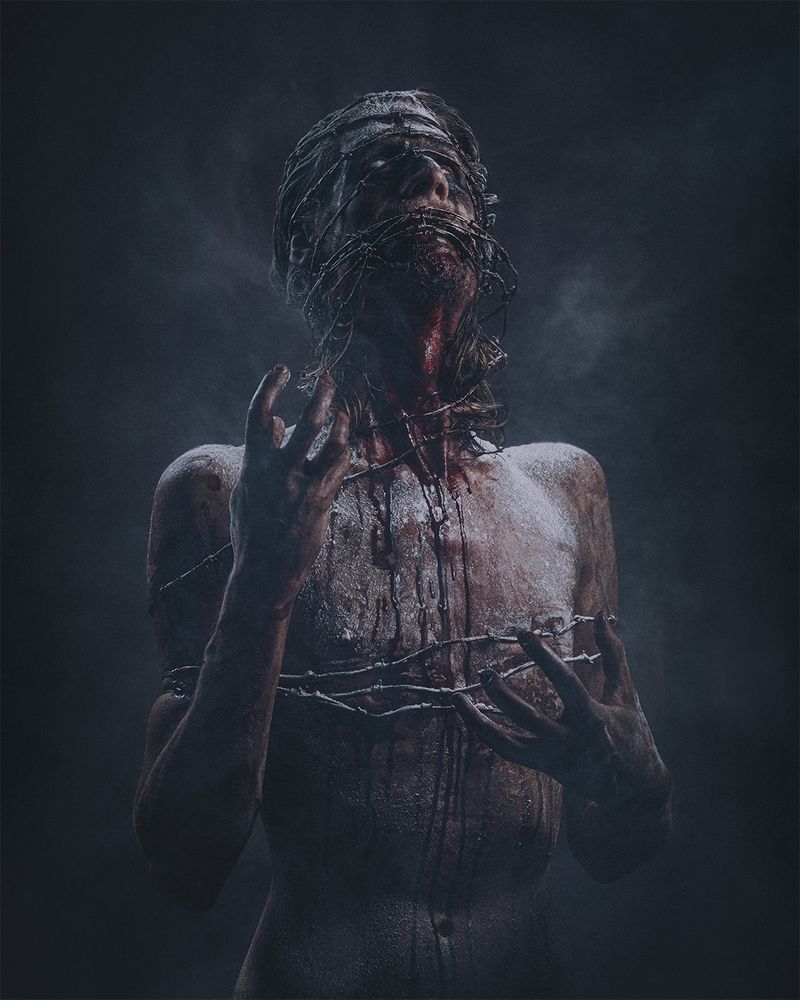 A photo of a naked figure wrapped in barbed wire and covered in blood, their eyes completely white. The air is filled with dust and haze.