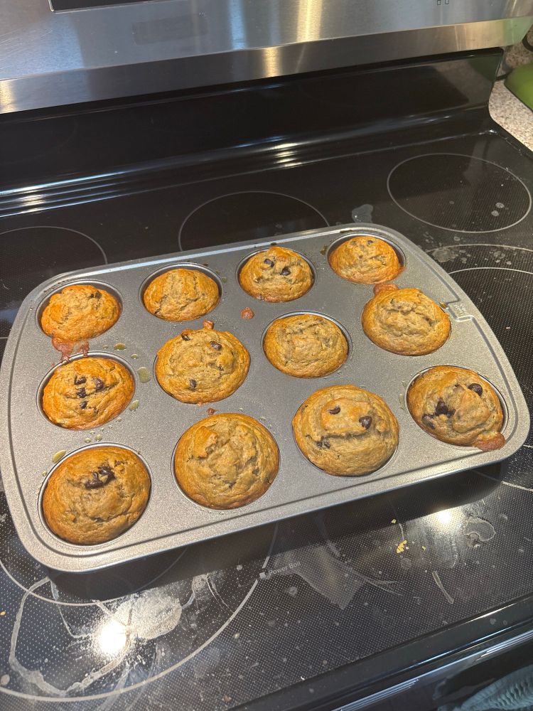 A muffin tray with 12 banana chocolate chip muffins on a dirty range top