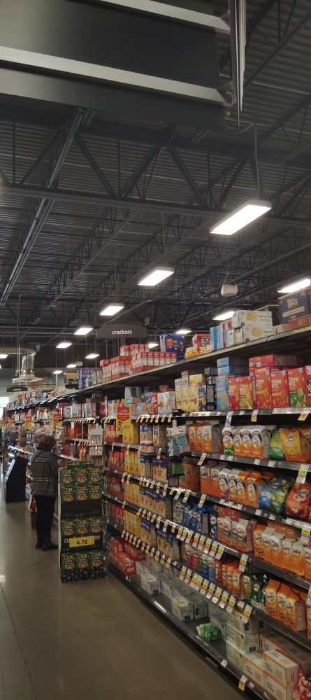 Photo of a grocery aisle containing shelves of various crackers and cookies. There is a sign above the shelves which says 'crackers', as well as another sign in the distance that says 'cookies'. Sitting atop the 'crackers' sign is a little sparrow which is looking to the right.