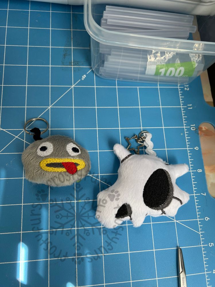 Two plush keychains lay on a blue fabric cutting board. 
The one on the left is the “Rock Facts” rock from Over the Garden Wall. It is made of grey minky with an embroidered face.
The one on the right is a Cubone skull from Pokemon. It is white minky with embroidered details.