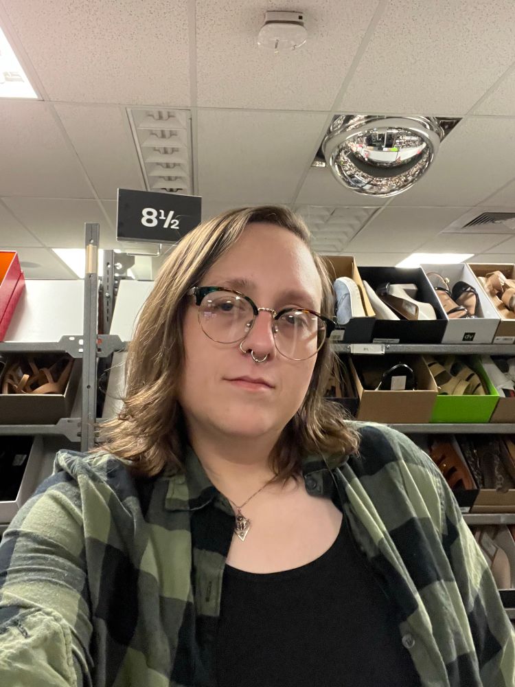 a selfie maya, a white, nearly middle-aged white trans woman with shoulder length brown hair, wearing a black tank top and a green and black plaid button up shirt, standing in the shoe isle of a retail store