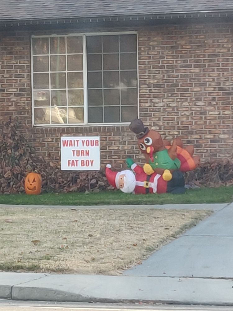 Someone's front yard. An inflatable turkey sitting on an inflatable Santa claus. A yard sign nearby says "wait your turn fat boy"