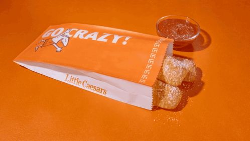 little caesars crazy bread breadsticks in an orange and white branded paper bag jext to marinara dipping sauce