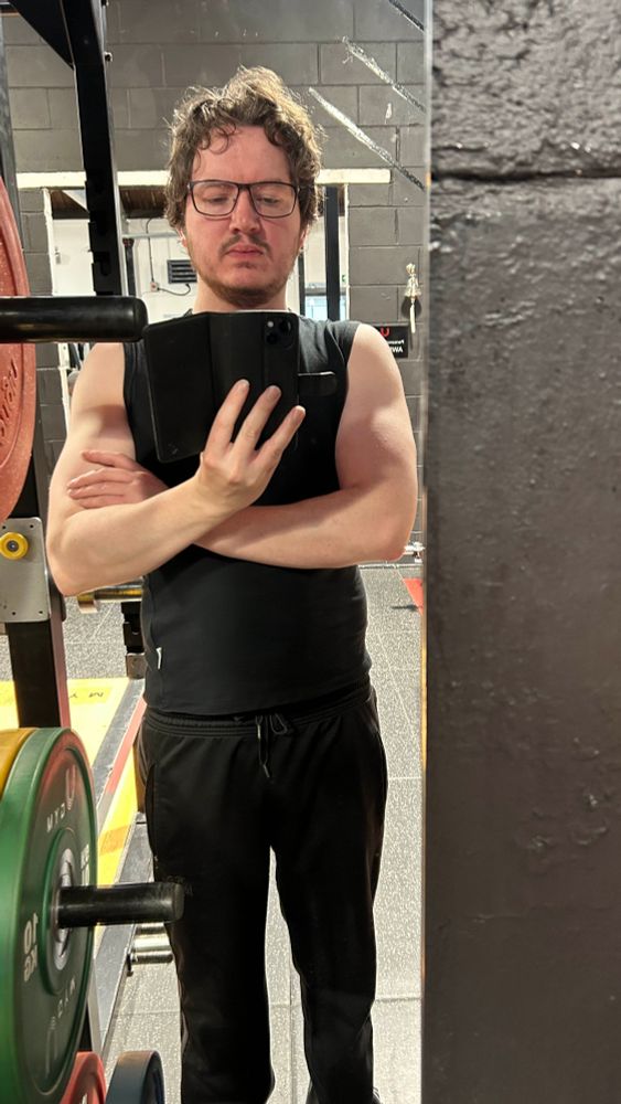 Man stands uncomfortably close to barbell rack to avoid disturbing anyone in gym. Black tank top and tracksuit bottoms. Arms folded. Three fingers up holding phone because he's a big fan of Wario. Glasses and hair.