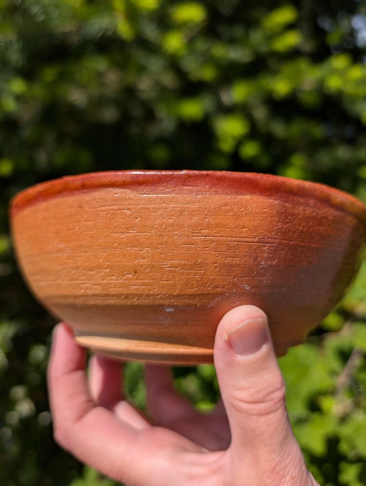 Hand holding for inspection a thick looking wood fired, orange tinted bowl. Sunny natural background