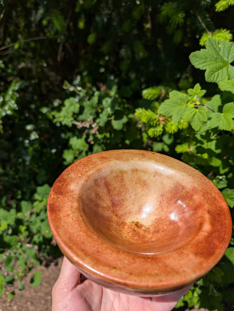 Hand holding for inspection a thick looking wood fired, orange tinted bowl. Sunny natural background