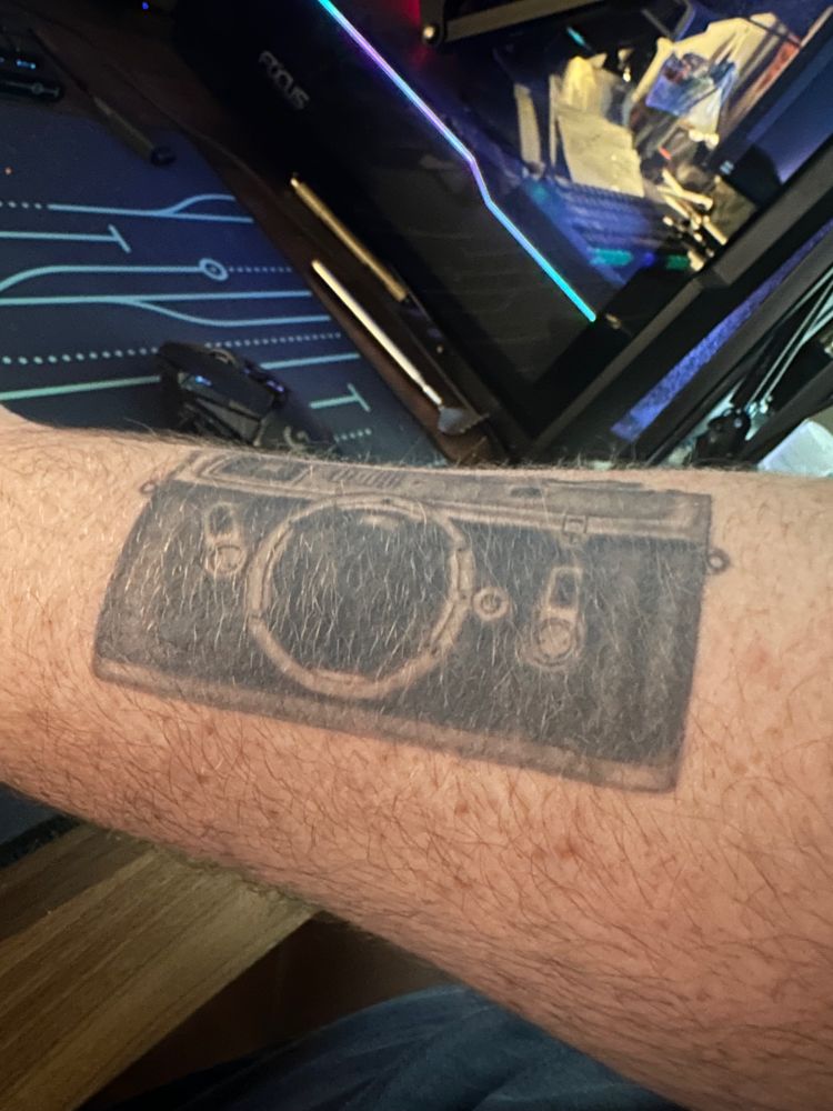 a picture of a white man's arm which has very faint freckles and a black and grey tattoo of a camera
