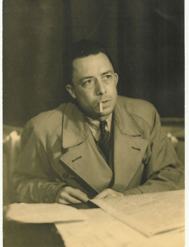 A sepia toned photo of French-Algerian philosopher Albert Camus wearing a trench coat and holding a cigarette in his lips.