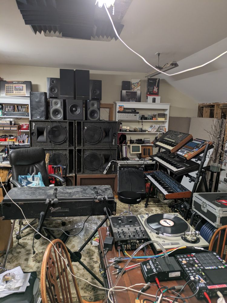 A somewhat chaotic-looking music production studio with DJ equipment, synthesizer keyboards, and other instruments and music gear haphazardly interconnected with cables to a large tower of disparate speakers.