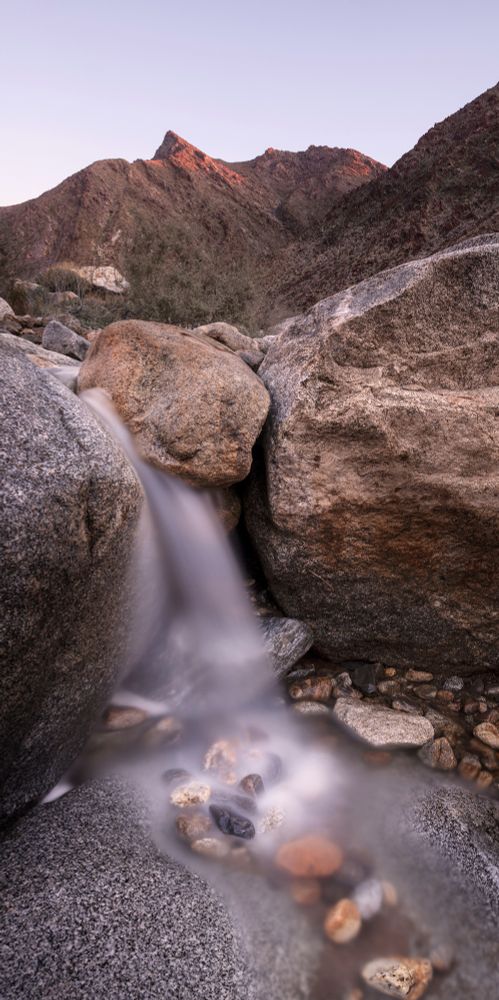 A photograph of a small waterfall in Palm Canyon, Anza Borrego Desert State Park.  Morning light strikes the tips of tall peaks rising over the canyon.