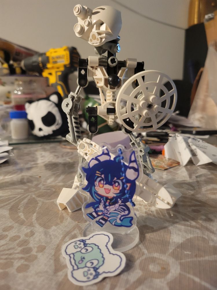 Photo of Litasea acrylic standee charm and mascot sticker in front of Kopaka Mata on a white, floral tablecloth in front of a pile of miscellaneous items including a dewalt drill, cat skull plushie, a lavendar circle, and some non-descript papers.