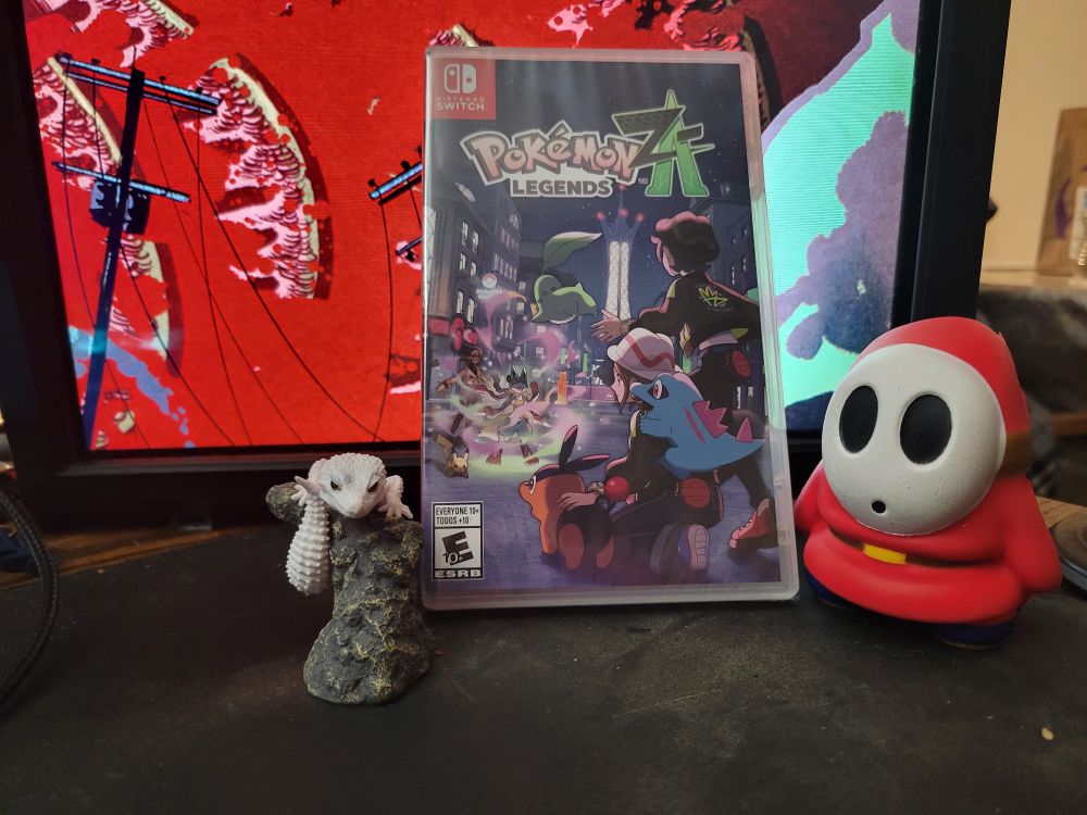 Physical copy of Pokemon Legends ZA for the Switch leaning on a vertical computer monitor showing the lower right side of a Shin Godzilla fan art by @mxmorgan on Cara. The game is flanked by a gachapon, albino gecko statue, and red Shy Guy from the Super Mario franchise made of foam.