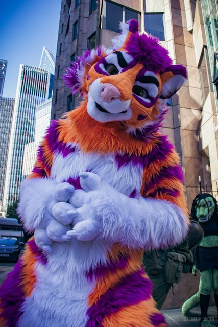 Arrimow, an orange, purple and white tiger fursuit, looking slightly down at the camera, his head cocked to the side. In the background there is a green dinosaur fursuiter and a lot of tall business type city buildings.