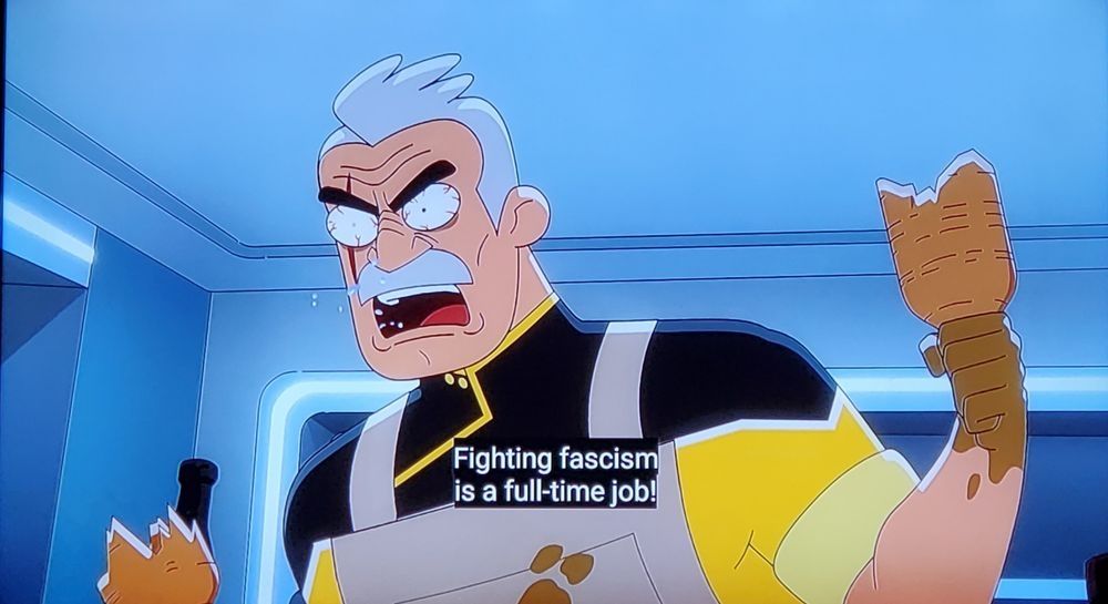 Our savior from fascism Shaxs, wearing overalls over his uniform, holds a broken bottle as a weapon in his left hand while he shouts "Fighting fascism is a full-time job!" 
