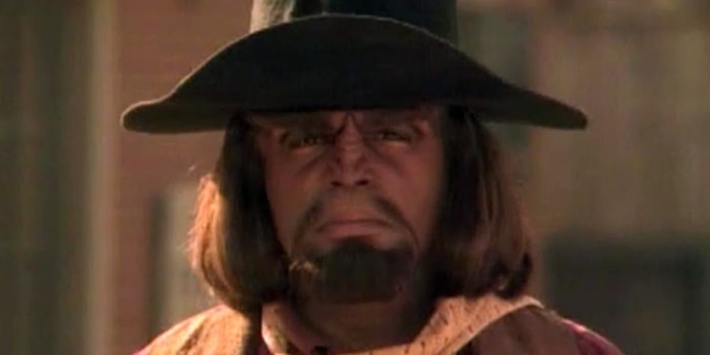 The scene is Star Trek. Onscreen is a Klingon whose name is Worf -- and he is not a merry man. No, he's wearing clothes that a gunfighter (foo fighter?) might wear. He's lit softly from his right, and he's wearing a large hat with a large brim. 🖖