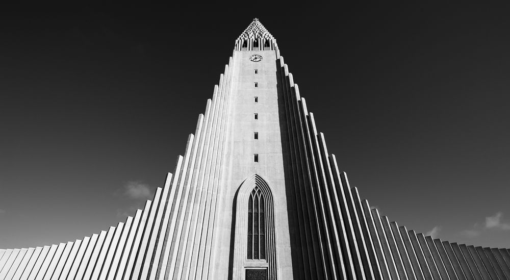 A striking black-and-white photograph of Hallgrímskirkja, the iconic Lutheran church in Reykjavík, Iceland. The image captures the church’s soaring, symmetrical concrete façade from a low angle, emphasizing its sweeping vertical lines that resemble basalt columns. The central tower rises sharply toward the sky, topped with a cross and a clock face, while the deep shadows and bright highlights accentuate the dramatic geometry of the structure against a dark, cloudless sky.