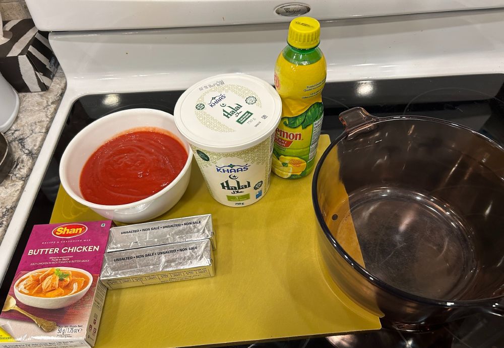 on a stovetop, a set of ingredient (strained tomatoes, plain yoghurt, lemon juice, butter chicken mix, two sticks of unsalted butter) and a pot