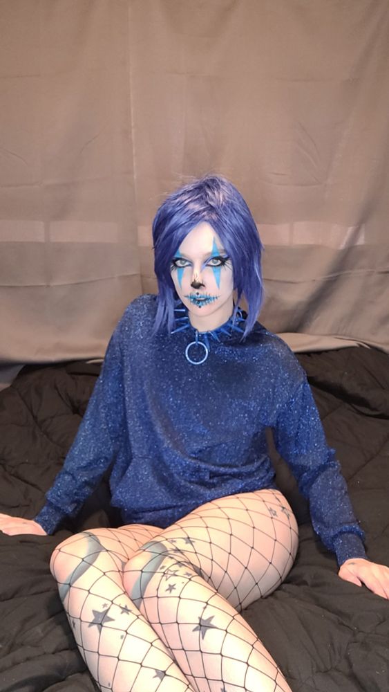 Blue haired clown with blue spooky makeup and blue sparkly shirt showing long legs wearing fishnet with doll joint and star tattoos.