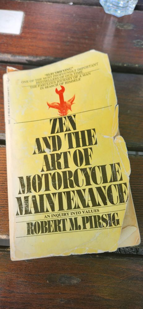 Photo of the cover of paperback novel "Zen and the Art of Motorcycle Maintenance", subtitled "An Inquiry into Values", by Robert M. Pirsig. Black text and red Illustration on faded & torn yellow cover.