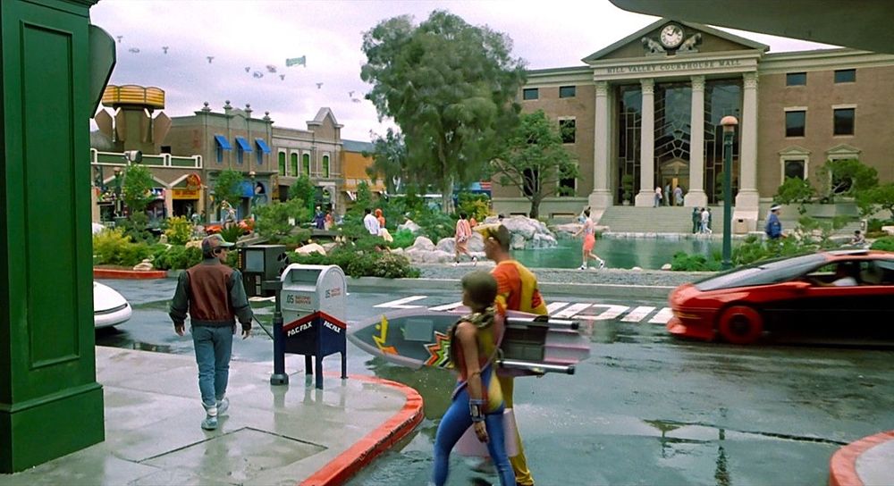 Still shot from a scene in Back To The Future 2. Marty McFly walks next to a mail dropbox as two pedestrians pass in the foreground. A red car is coming in from the right of the frame. The courthouse with the clock tower is present in the background.