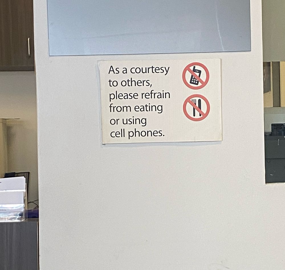 A sign on a plain white wall that says: "as a courtesy to others, please refrain from eating or using cell phones."