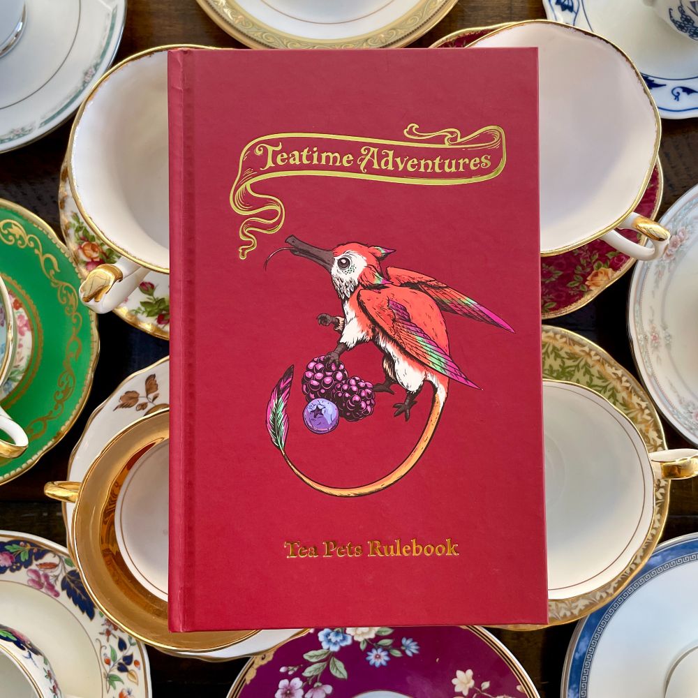 Teatime Adventures Tea Pets Book (red with a flower dragon centered on the cover) sits atop a table filled with teacups