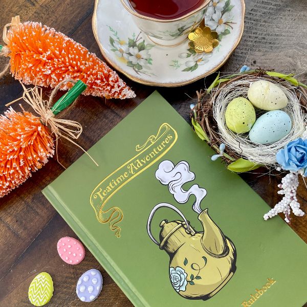 a green Teatime Adventures 2E core rulebook sits on a wooden table alongside a cup of tea, and easter decorations 