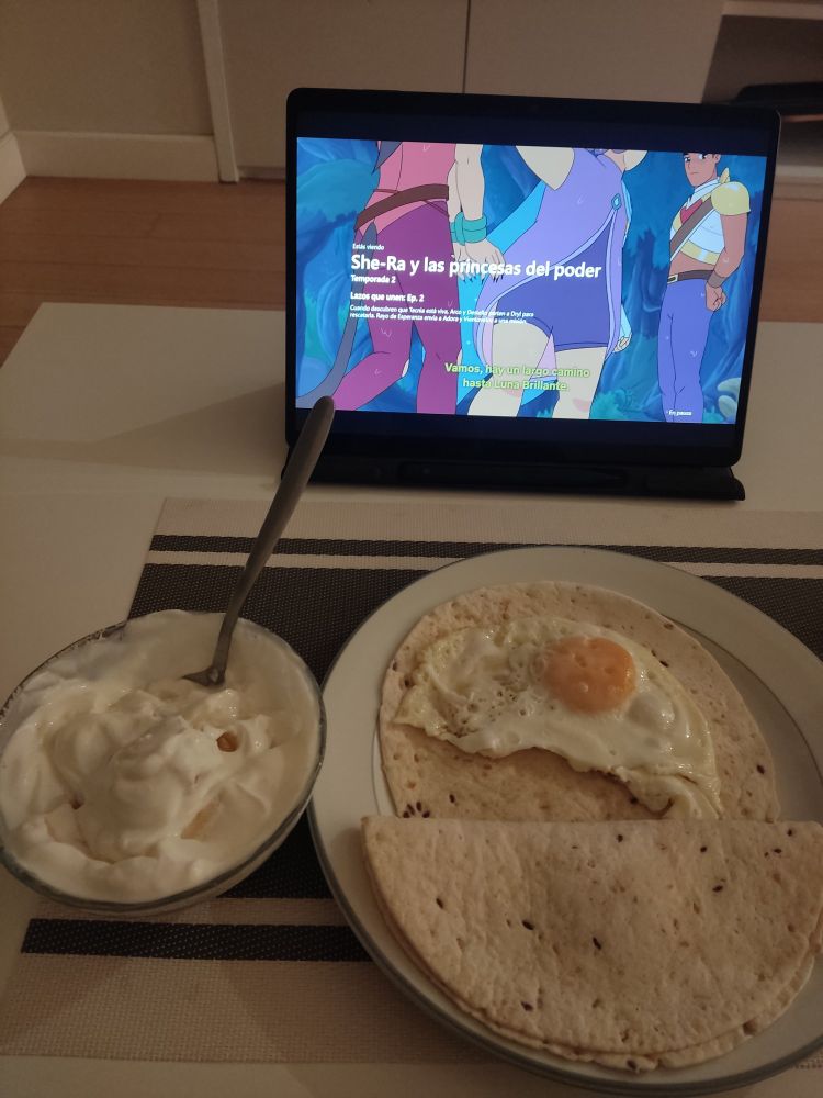 Se ve, en un plato, un huevo frito en una tortilla de maíz, y una segunda tortilla doblada ocultando el segundo huevo. También un yogurt con un plátano machacado revuelto en el mismo, en un cuenco de cristal. Al fondo se ve una escena de She-Ra, Temporada 2 Ep.2 Lazos que unen, donde salen Catra, con las manos atadas, y Glimmer agarrándola