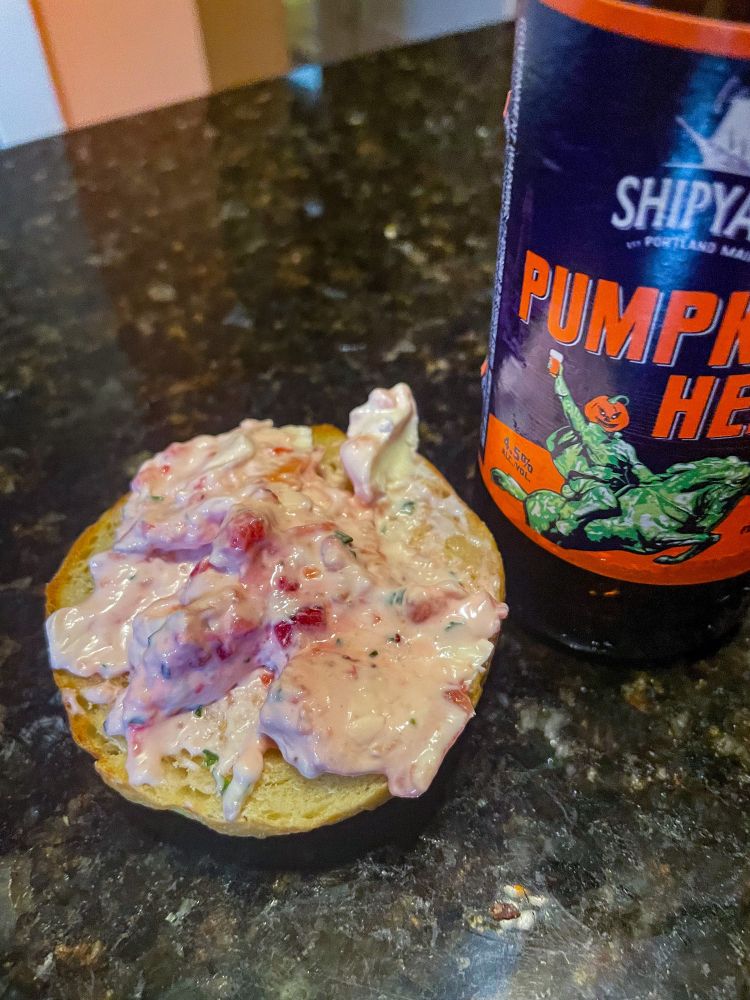 A half of a bagel with strawberry basil cream cheese sitting on a marble table top beside a Shipyard Pumpkinhead beer.