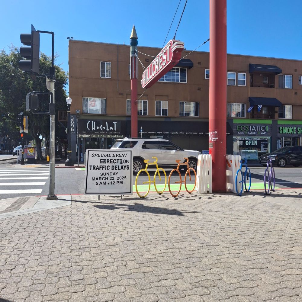 Someone has defaced a sign to say Erection Traffic delays instead of Expect Traffic Delays. The sign is below the Hillcrest neighborhood sign next to a rainbow bike rack. 