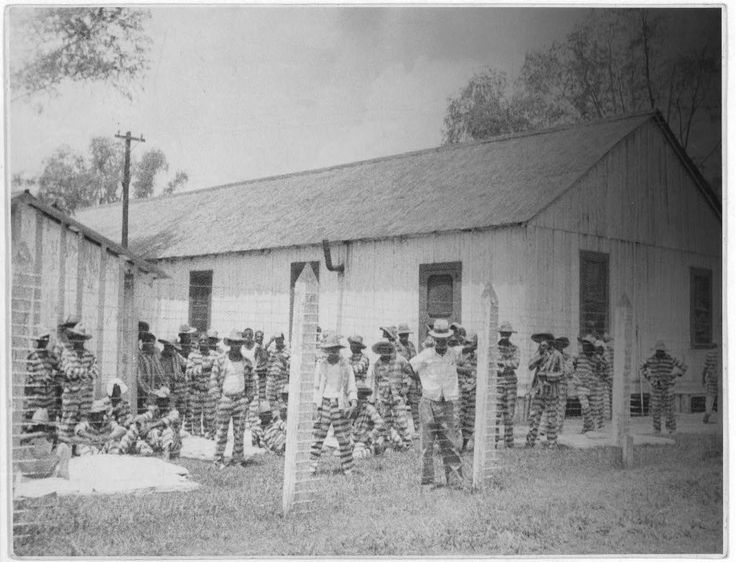 Men in prison stripes stand behind tall barbed wire fence, in front of a low building. Most wear hats in the summer sun. 