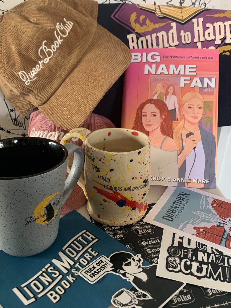 A still life image of the book BIG NAME FAN with baseball hats that read "Queer Book Club" and "Romance Reader," a Starry Night cat cafe mug, the logo of Lion's Mouth Bookstore, a mug that says "imagine being afraid of books and dragqueens," a Bound to Happen Books totebag, and stickers that say "protect trans kids" and "protect trans folks," as well as "f*** off fascist" and "f*** off Nazi scum."