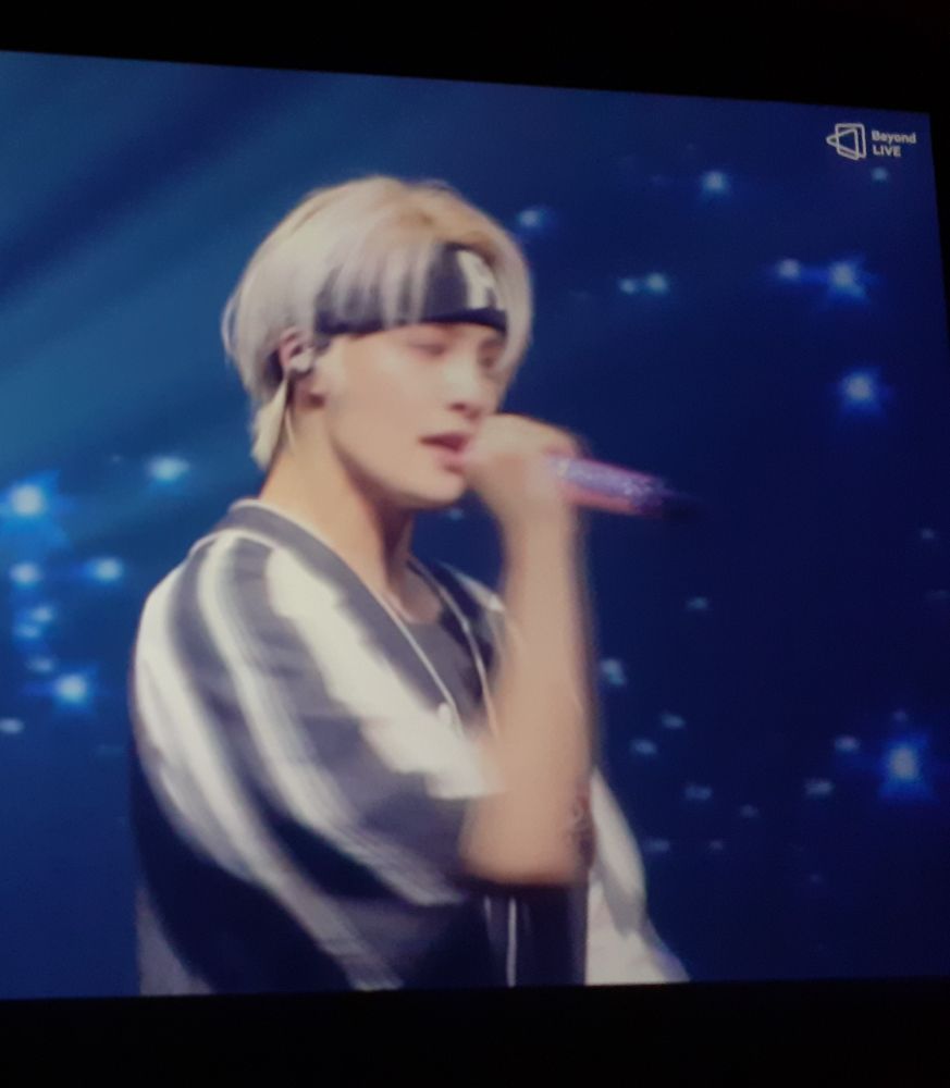 Blurred photo of computer screen with Yangyang of wayv holding a microphone and singing. His floppy white-blond fringe is held away from his head by a thick black headband