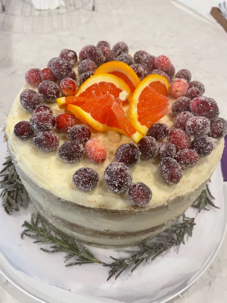 A holiday cake with three gingerbread cake layers, cream cheese frosting, and cranberry sauce filling. It is decorated as a ‘naked’ cake with sugared cranberries, rosemary, and orange slices. 