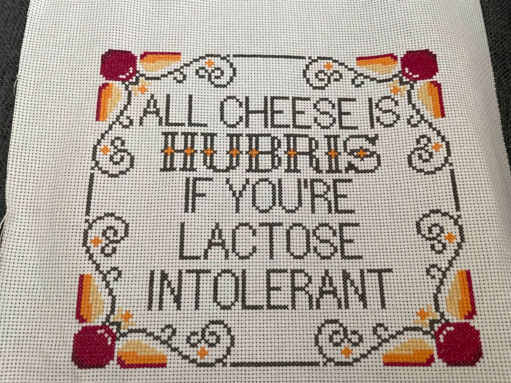 A completed cross-stitch featuring wheels and slices of cheese sealed in red wax, surrounding text reading 'All cheese is hubris if you're lactose intolerant'. 'Hubris' is pronounced 'Hubrie' for this joke.