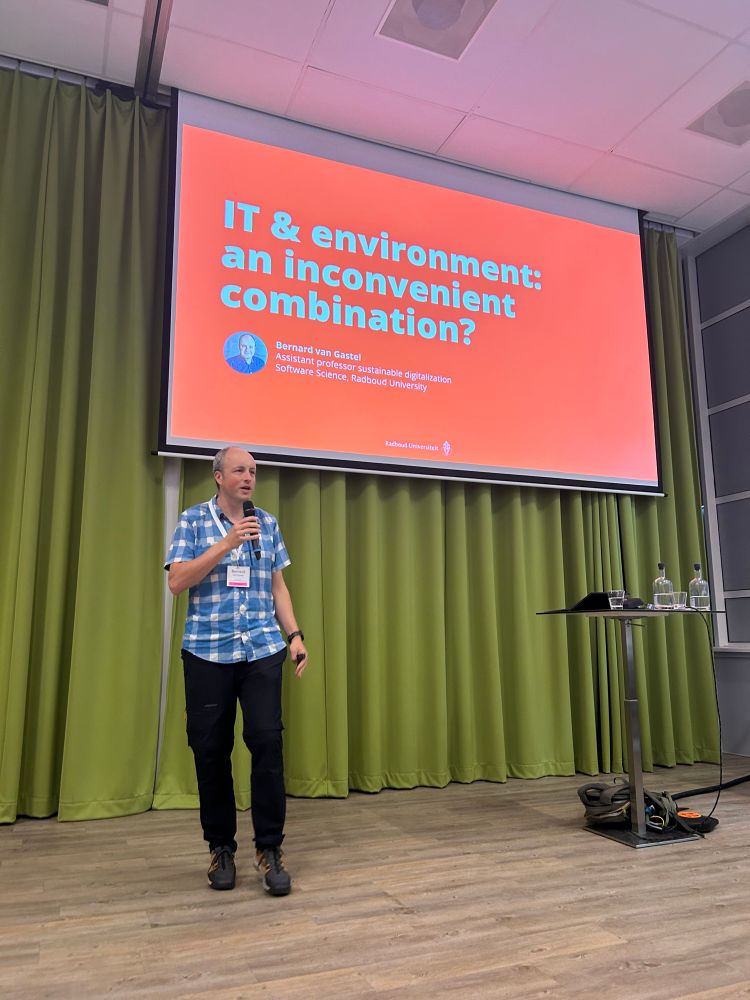 Links op de foto Bernard van Gastel. Hij draagt een zwarte broek en een blauw witte blouse met korte mouwen. Achter hem een heel groot scherm. Op het scherm de tekst: IT & Environment an inconvenient combination. 