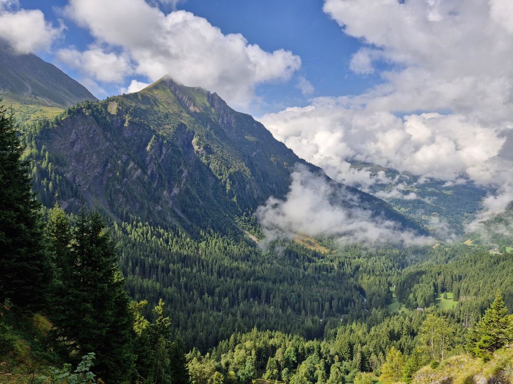 A picture of a verdant valley covered in trees. In the upper right quadrant, a peak can be seen. There are a lot of clouds, but the weather is sunny and some blue parts of the sky can be seen.