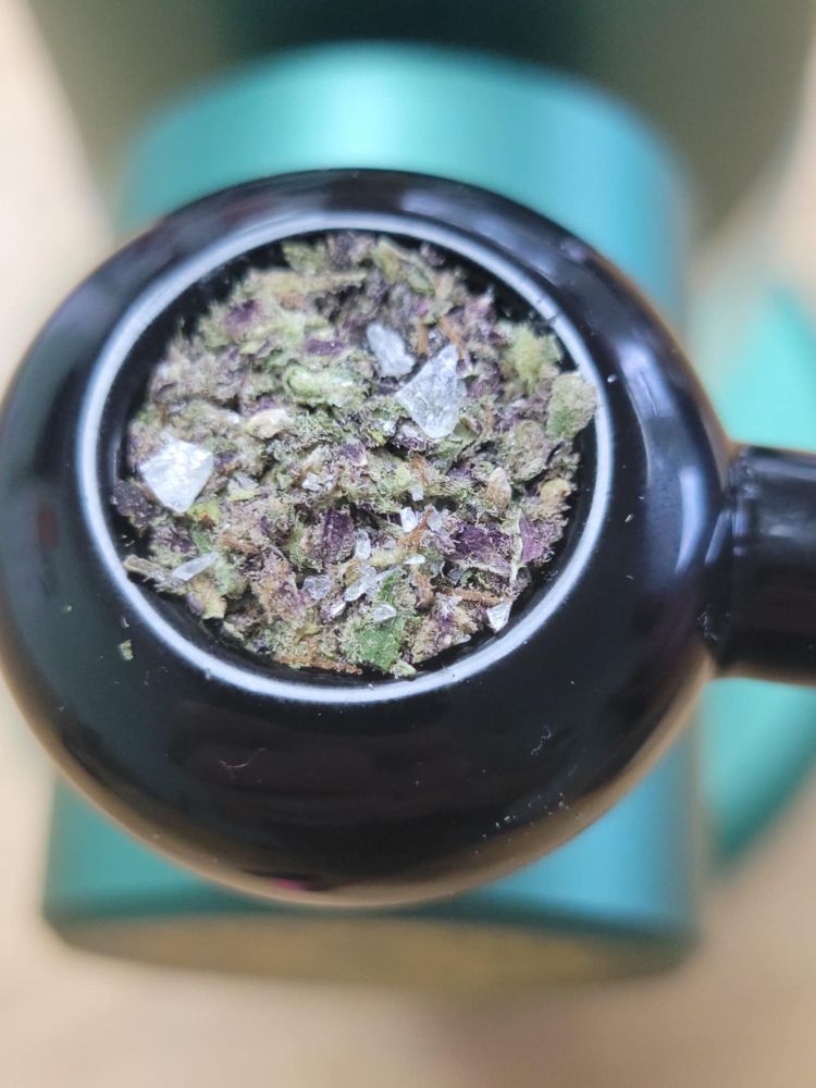 A packed bowl of cannabis. It has green and lots of purple tones with a white diamond concentrate on top