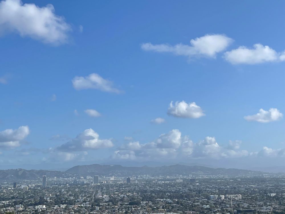 a view of a part of Los Angeles from above. City, trees, blue sky, puffy clouds here and there. In the background are mountains and if zooming in one can see the Griffith Park Observatory. 
