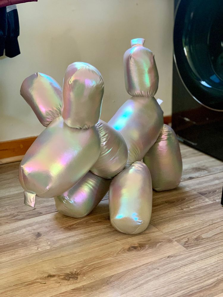 Picture is of silver + iridescent balloon dog plush, on wood textured floor, with white wall, and washing machine in background. 